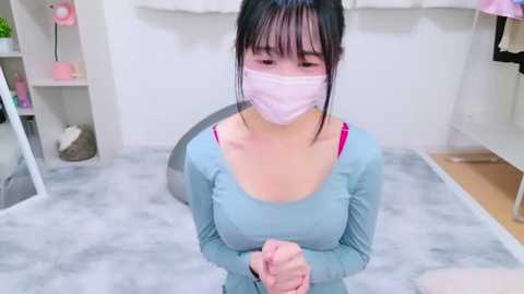 Media: Video of an East Asian woman with dark hair and pale skin, wearing a pink mask and light blue top, kneeling on a gray carpet, looking distressed in a modern, minimalist room.