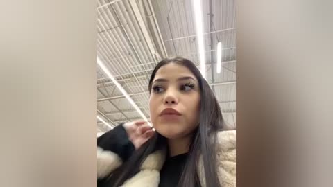 Media: Video of a young Latina woman with long, straight dark hair, wearing a black top and a fur coat, in an industrial setting with white metal beams and fluorescent lights.