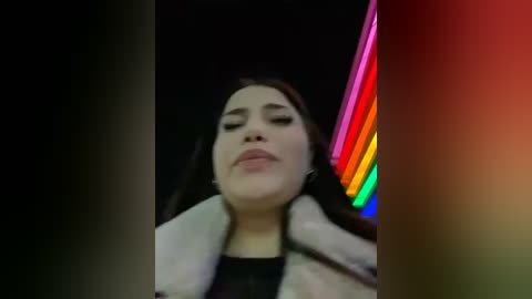 Media: A low-resolution video of a woman with fair skin, dark hair, and a neutral expression, wearing a fur-lined coat, set against a neon-lit background with vibrant colors.