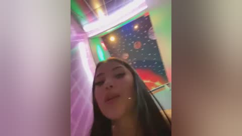 Media: A video of a young Asian woman with long black hair, looking slightly downwards, in a dimly lit, colorful room with neon lights, a starry ceiling, and a red object in the background.