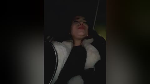 Media: Video of a young woman with light skin, dressed in a black jacket with white fur trim, lying on a dimly lit floor, her head turned to the side, eyes closed, and mouth slightly open.