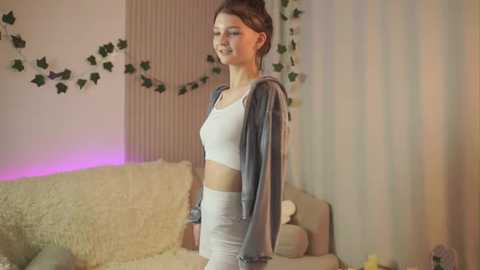 Media: Video of a young woman with light skin and brown hair, wearing a white crop top and high-waisted pants, standing in a cozy living room with soft lighting and green ivy decor.