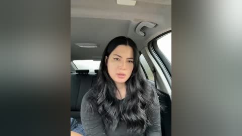 Media: Video of a young woman with long, wavy black hair, light skin, and a full face of makeup, wearing a black top, seated in a car.