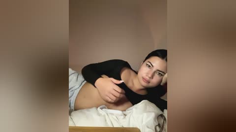 Media: Video of a young woman with olive skin, long black hair, lying on a bed in a black long-sleeve top, covering her breasts, with a hand over her stomach.