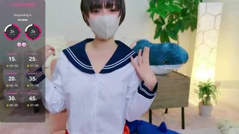 Media: Video of an Asian woman with short dark hair wearing a white sailor school uniform with a navy blue collar and face mask, standing in a modern room with a green plant, blue plush toy, and a wooden table.