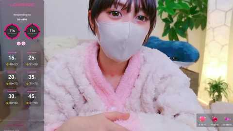 Media: A video of an Asian woman in a fluffy pink robe, wearing a white face mask, sitting indoors. The background shows a potted plant and a blue cushion.