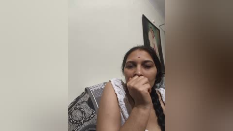 Media: Video of a South Asian woman with long black hair, wearing a white and grey patterned sari, covering her mouth with her hand, in a plain white room with a framed picture on the wall.