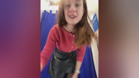 Media: Video of a smiling Caucasian woman with long brown hair, wearing a red sweater and black skirt, standing in a blue curtained room.