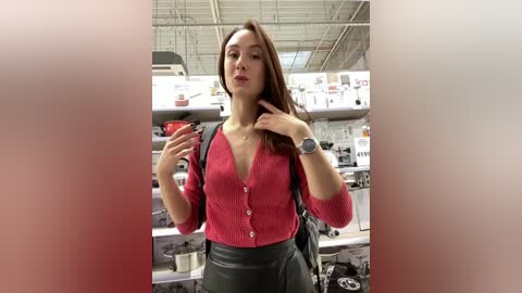 Media: Video of a young woman with long brown hair, fair skin, and medium build, wearing a red cardigan and gray pants, standing in a brightly lit kitchen store. She poses with a neutral expression.