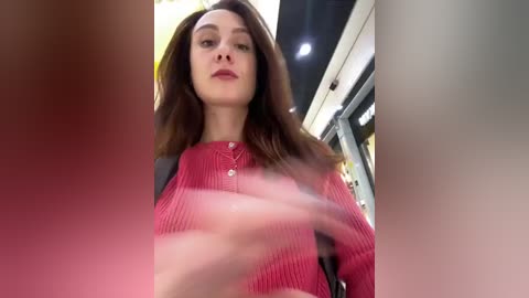 Media: Video of a young woman with straight, shoulder-length brown hair, wearing a red, long-sleeved, ribbed knit top. She gazes downward, her face slightly blurred. Background shows a modern indoor setting with overhead lighting and glass structures.