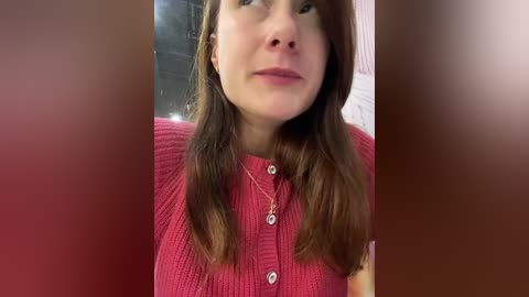 Media: Video of a fair-skinned woman with shoulder-length brown hair, wearing a red, textured, button-up cardigan, and a delicate necklace. Background is blurred, suggesting an indoor setting.