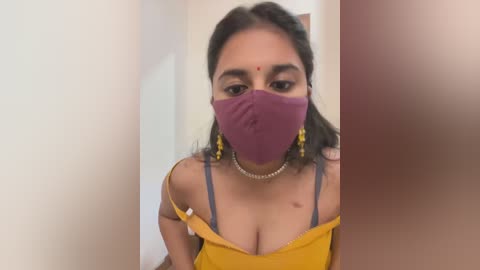 Media: A video of a young woman with medium brown skin, wearing a yellow off-shoulder top, purple mask, and yellow earrings. She has a small, red bindi on her forehead.