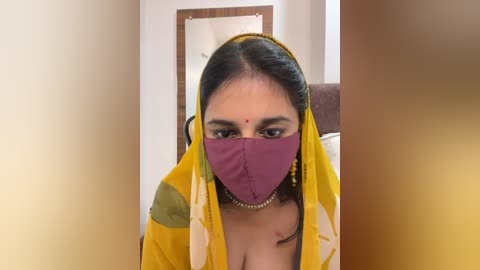 Media: Video of a woman with medium skin tone, dark hair, and wearing a yellow and green saree with a purple mask. She sits indoors, near a wooden door.