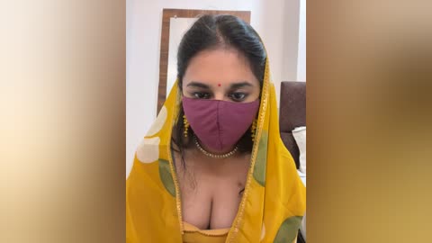 Media: Video of a South Asian woman with medium skin tone, wearing a yellow sari with green and white floral patterns, a purple mask, and a gold necklace, seated indoors.