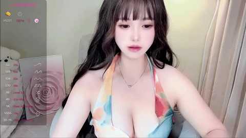 Media: Video of a young East Asian woman with long black hair, wearing a colorful tie-dye bikini top, indoors with soft lighting, blurred background.