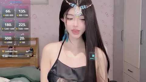 Media: Video of a young East Asian woman with long black hair, wearing a black satin dress and blue feather headdress, in a bedroom with pink wallpaper.