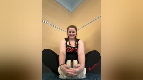 Media: Video of a smiling woman in a black tank top and leggings, sitting cross-legged on a blue carpeted floor, stretching her legs in a gym setting with beige walls.