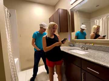 Media: Video of a blonde woman in a black tank top and red shorts texting while a man in a turquoise shirt and cap looks at a mirror in a modern bathroom with beige walls and dark wood cabinets.