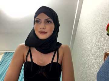 Media: Video of a light-skinned woman with a dark complexion, wearing a black headscarf and a black velvet dress with thin straps. She stands in a bathroom with a textured white wall and teal curtains in the background.