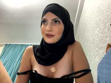 Media: Video of a woman with medium skin tone, wearing a black hijab and off-shoulder black top, with a floral pendant necklace. She has dark, expressive eyes and a subtle smile. Background includes white walls and teal curtains.