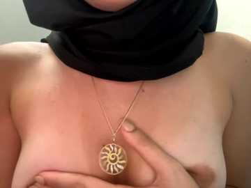 Media: Video of a topless woman with light skin, covering her breasts with her hands, wearing a black headscarf and a gold necklace with a sun-shaped pendant.