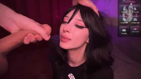 Media: A video of a pale-skinned woman with black hair, wearing a black top, being held by the head by a hand, in a dimly lit room with purple and red tones.