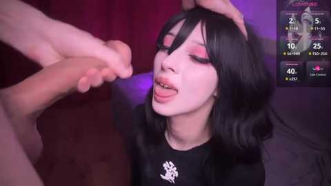 Media: Video of a pale-skinned woman with long black hair, wearing black makeup, kneeling, licking a man's hand. Background has a red curtain and a purple light.