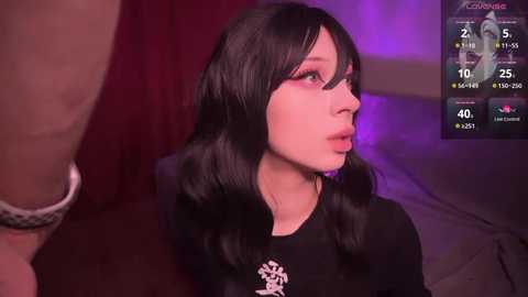 Media: A video of a pale-skinned, slender young woman with long black hair and dramatic makeup, wearing a black sweater. Background features a purple-lit room with a \"Love Island\" TV show overlay.