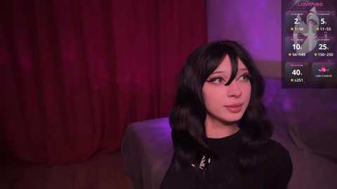 Media: Video of a young woman with long black hair, pale skin, and large breasts, wearing a black top, sitting on a bed with red curtains, illuminated by purple light.