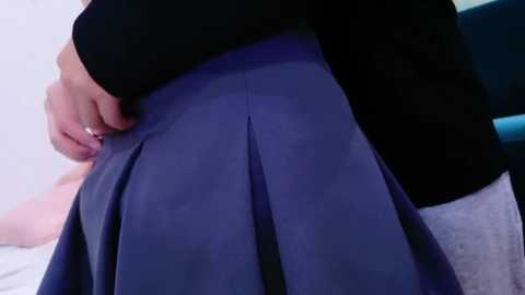 Media: Video of a woman's hand adjusting the hem of a pleated, dark blue skirt over a light gray top. The background is blurred, featuring a teal chair.