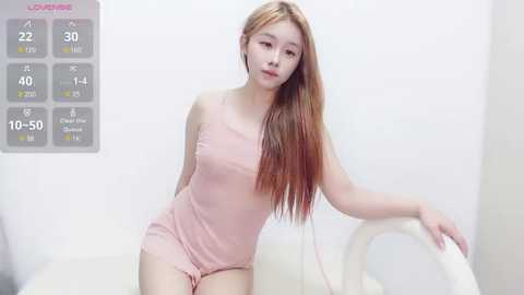 Media: Video of a young Asian woman with long, straight, light brown hair, wearing a light pink, form-fitting dress, leaning on a white chair in a bright, minimalist room.