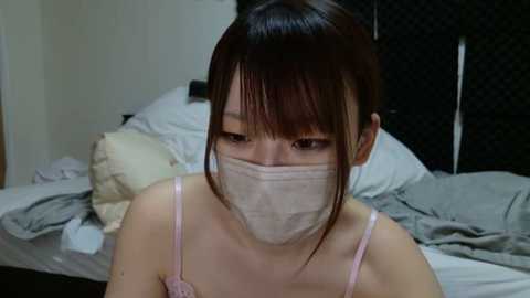 Media: Video of an Asian woman with straight black hair, wearing a pink camisole, light beige mask, and a light gray face shield, seated on a bed with messy gray sheets.