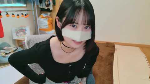 Media: Video of a young Asian woman with short black hair, wearing a black top, beige pants, and a white bandage over her mouth. She sits on a brown sofa in a cluttered room with a white wall and a messy shelf in the background.