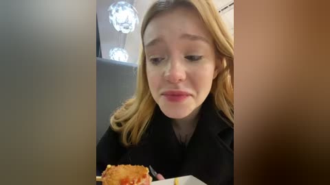 Media: Video of a young woman with light skin and long blonde hair, wearing a black coat, eating a hot dog with ketchup inside a restaurant with modern, circular light fixtures on the wall.