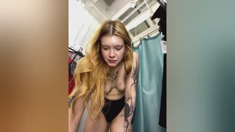 Media: Video of a topless, tattooed young woman with long, wavy blonde hair in a dimly lit, messy bedroom with clothes and a teal curtain in the background.