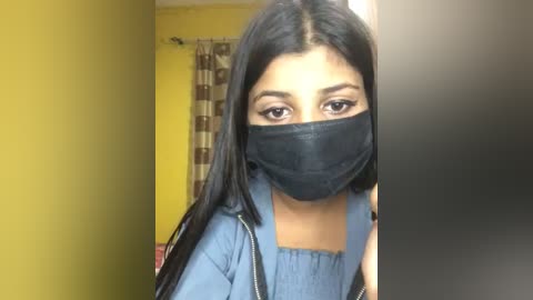 Media: Video of a young woman with medium-brown skin, long black hair, and dark eyes, wearing a black face mask, blue top, and black jacket, in a room with yellow walls and a striped curtain.