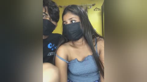 Media: Video of a young couple wearing black face masks, with the woman in a blue off-shoulder top and the man in a black shirt. Background shows a yellow wall and a partially visible sign.