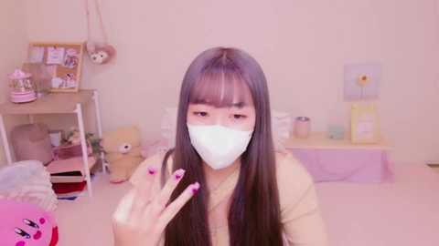 Media: Video of an Asian woman with long black hair, wearing a white face mask, beige top, and pink manicured nails, in a pastel-themed, tidy bedroom with stuffed animals, shelves, and floral decorations.