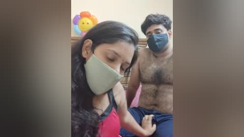 Media: A video of a South Asian woman with long black hair and a mask, seated on a pink couch, next to a shirtless man with a beard, wearing a mask. A colorful toy is visible in the background.