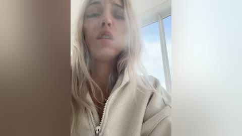 Media: A video of a young, fair-skinned woman with long, platinum blonde hair, wearing a beige hoodie, with her eyes closed and mouth slightly open, taken indoors near a large window with natural light streaming in.