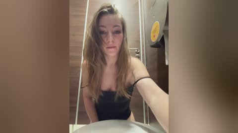 Media: Video of a young Caucasian woman with long, light brown hair, wearing a black strapless top, taking a selfie in a public restroom with a wooden door and a yellow sticker on it.