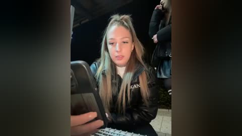 Media: Video of a young blonde woman with long hair, wearing a black jacket, seated indoors, holding a smartphone, and looking at the camera.