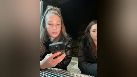 Media: Video of a young woman with long blonde hair in a ponytail, wearing a black leather jacket, taking a selfie with a smartphone at night, outdoors, with blurred background.