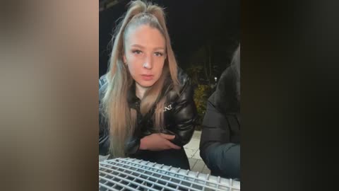 Media: Video of a young woman with long blonde hair tied in a high ponytail, wearing a black leather jacket, seated on a tiled floor, next to a white keyboard. Background is dimly lit, suggesting an indoor, possibly nighttime setting.