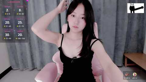 Media: Video of a young Asian woman with long black hair, wearing a black spaghetti-strap top, sitting on a pink chair in a room with grey curtains.