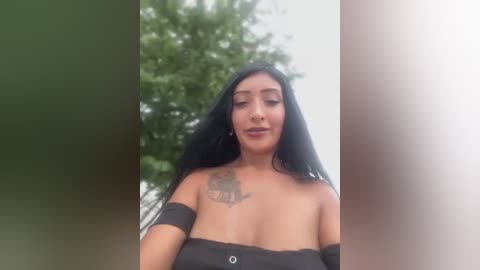 Media: Video of a young woman with long black hair, medium skin tone, and a tattoo on her shoulder, wearing a strapless black top. Background shows blurred green foliage and a cloudy sky.