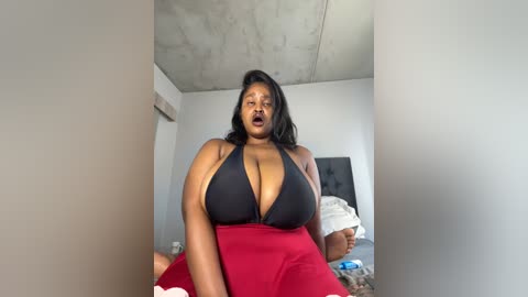 Media: Video of a plus-size Black woman with medium-dark skin tone, wearing a black halter top and red skirt, sitting on a bed in a minimalistic room with exposed concrete ceiling and plain walls.