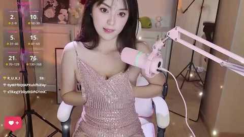 Media: Video of an Asian woman with long black hair, wearing a pink sequin dress, sitting in a chair with a microphone, in a home studio with soft lighting and floral decor.