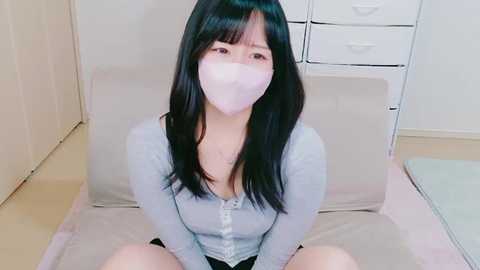 Media: Video of an East Asian woman with long black hair and light skin, wearing a light gray long-sleeve shirt, black shorts, and a white face mask, kneeling on a pink cushion in a simple, minimalistic room with white cabinets and drawers.