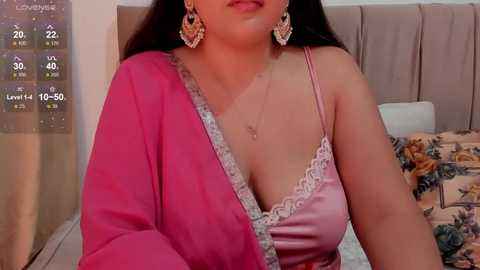 Media: Video of a South Asian woman in a pink, lace-trimmed top, wearing large gold earrings, in a bedroom with a floral bedspread.